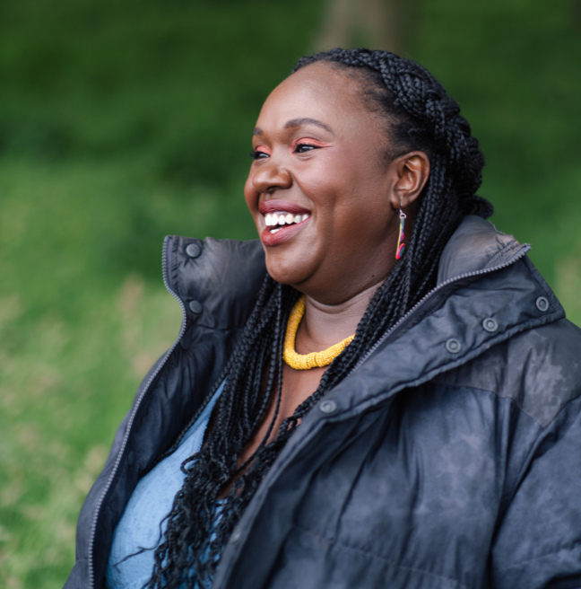 A black woman, with braided hair, wearing a navy coat over a blue denim dress.  She's bright jewellery on, necklace and earrings, and is smiling and laughing off into the distance.