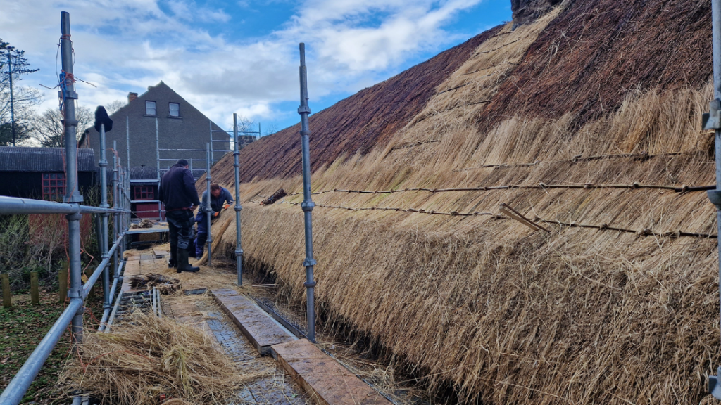 A man stands on scaffolding set up along a cottage. He is in the middle of rethatching the roof.