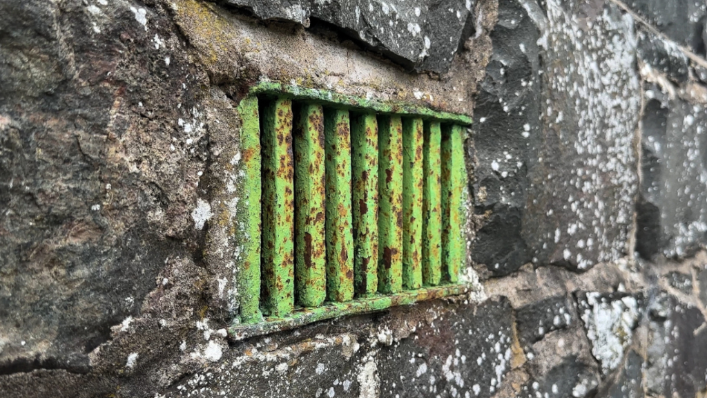 A green grate embedded into the side of a stone wall, intended to air out the building for damp.