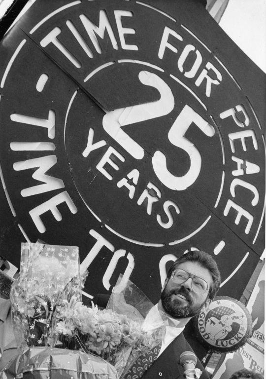 Black and white image of Gerry Adams standing in front of mural 'Time for peace...'