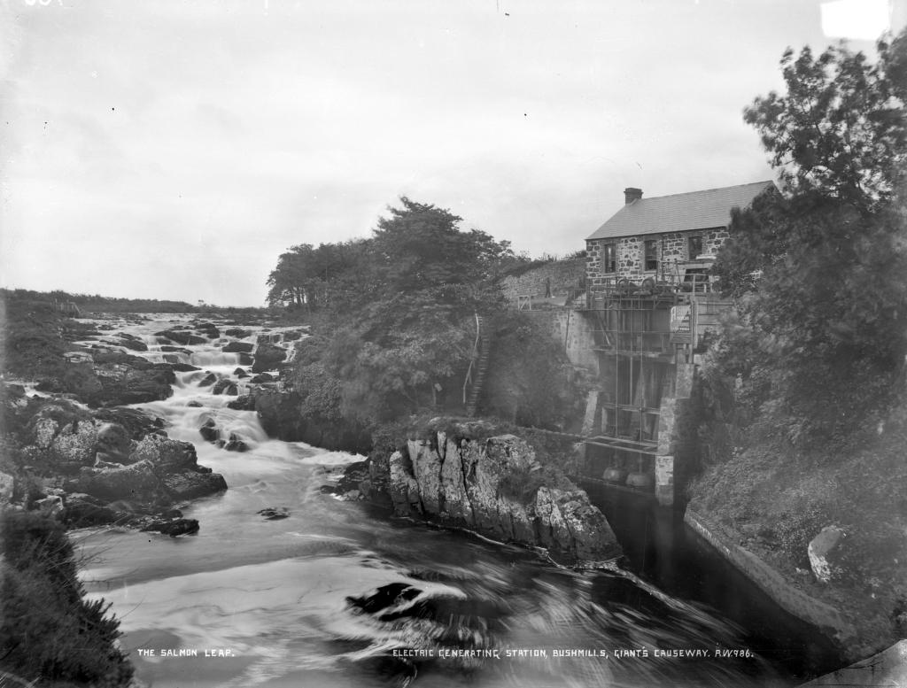 A black and white photograph of a wide river. On the left, a series of small waterfalls are identified as a salmon run. On the right is a hydroelectric station, consisting of a small stone cottage at the top of a steep hill, with a contraption stretching below to the river.