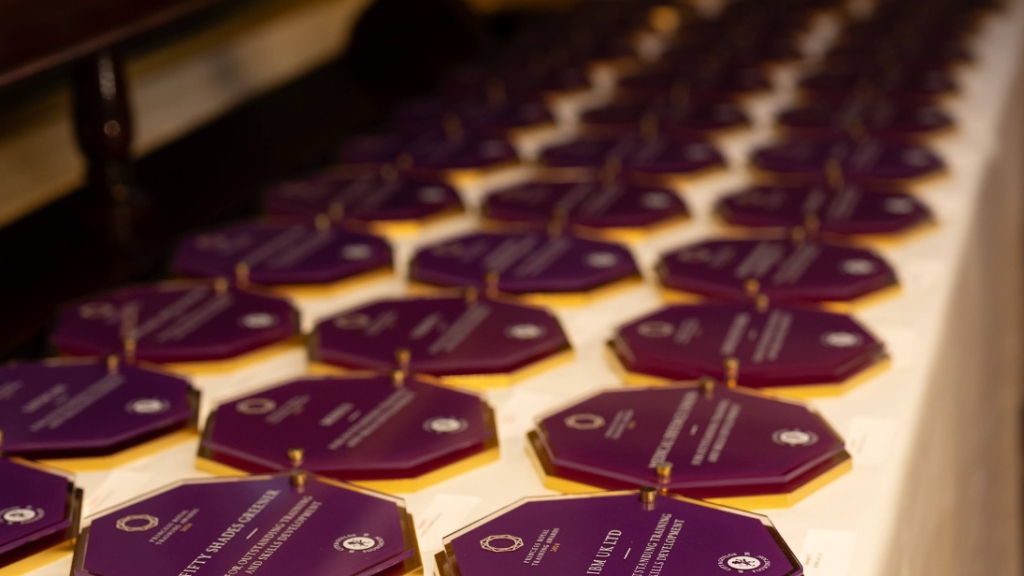 A table with a white tablecloth on it, displaying three rows of awards in a purple and gold hexagon design. They are the Princess Royal Training Awards.