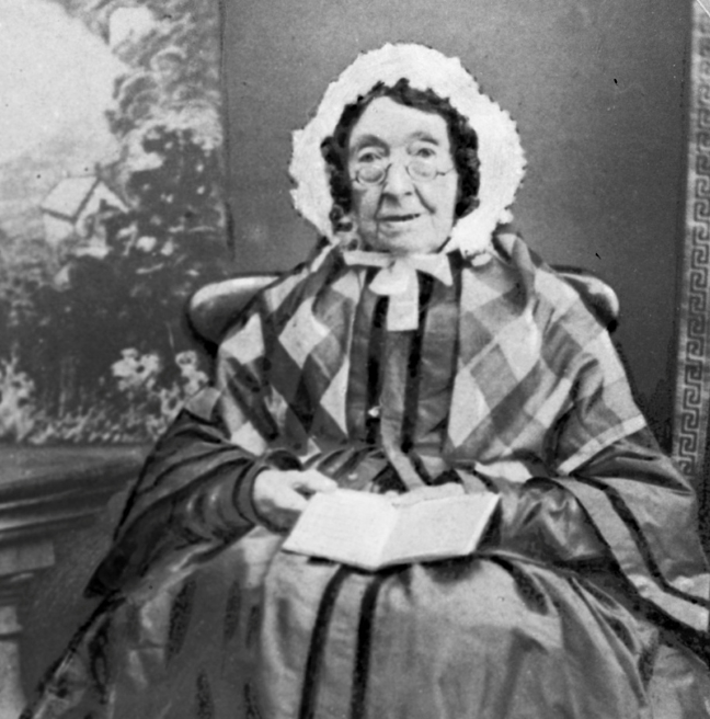 An older woman sits for a black and white photograph. She is dressed in typical 1800s fashion., with a book open on her lap.