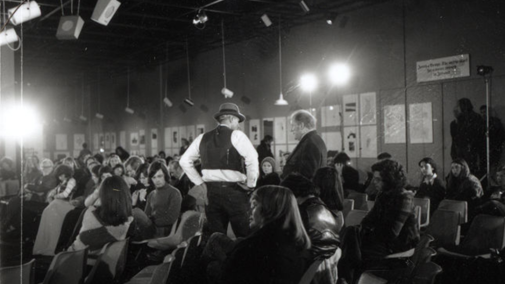 A black and white photograph of Joseph Beuys in the middle of an audience at his lecture. 