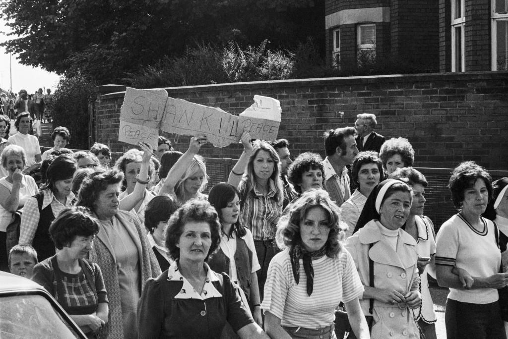 A black and white photo of a march for peace. Three women in the crowd hold up a handmade sign reading 'Shankill peace'.