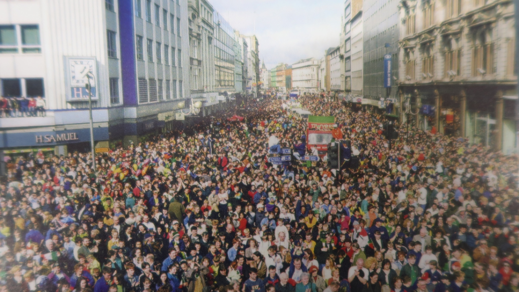 Postcard of photograph of Belfast's first ever city centre St Patrick's Day Carnival Parade, March 17 1997. The streets are full of people, with some climbing buildings and poles.