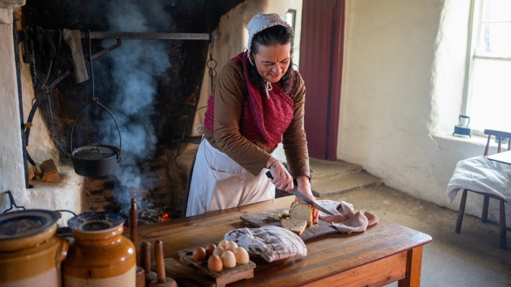 A woman dressed in traditional 19th century clothing stands in a cottage cutting bread.