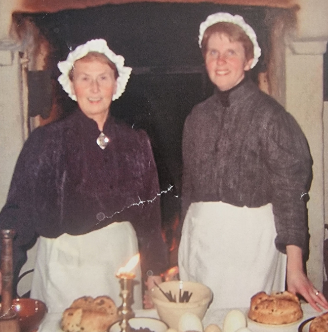 Two woman stand in a cottage dressed in costume. The costumes are traditional women's clothing from the 1800s.