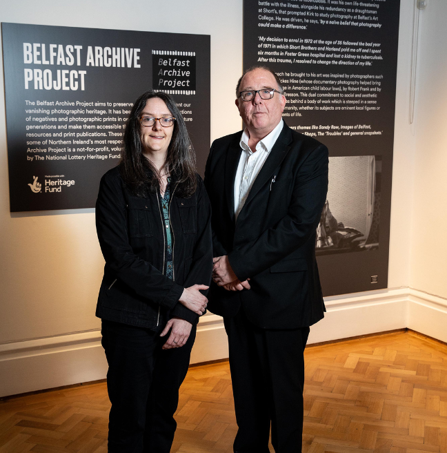 A photo of a man and a woman standing in front of a black and white photographic exhibition display. Both are smartly dressed in black, smiling at the camera, wearing glasses and have their hands crossed in front of them.