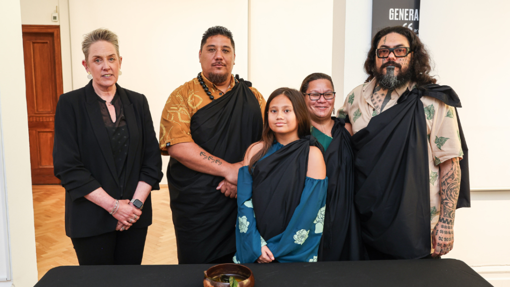 A group of five people standing in front of a table with a black table cloth, all wearing either black or traditional dress, during a repatriation ceremony at Ulster Museum.