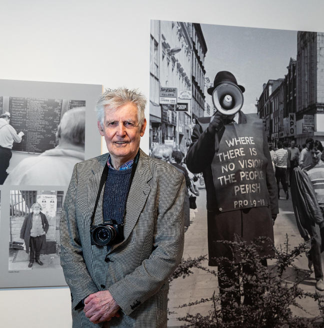 A photo of a man dressed in a grey suit with navy jumper, grey/white hair and a camera hanging around this neck. He is a photographer and he is standing in front of a black and white panel featuring one of his photographs.st Archive Project' with a blurb of white text on a black background plus National Lottery Heritage Fund logo and Belfast Archive Project logo.