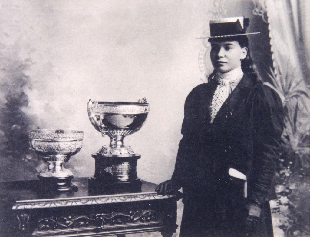 A black and white photograph of a woman wearing era-appropriate clothes standing next to two trophies.