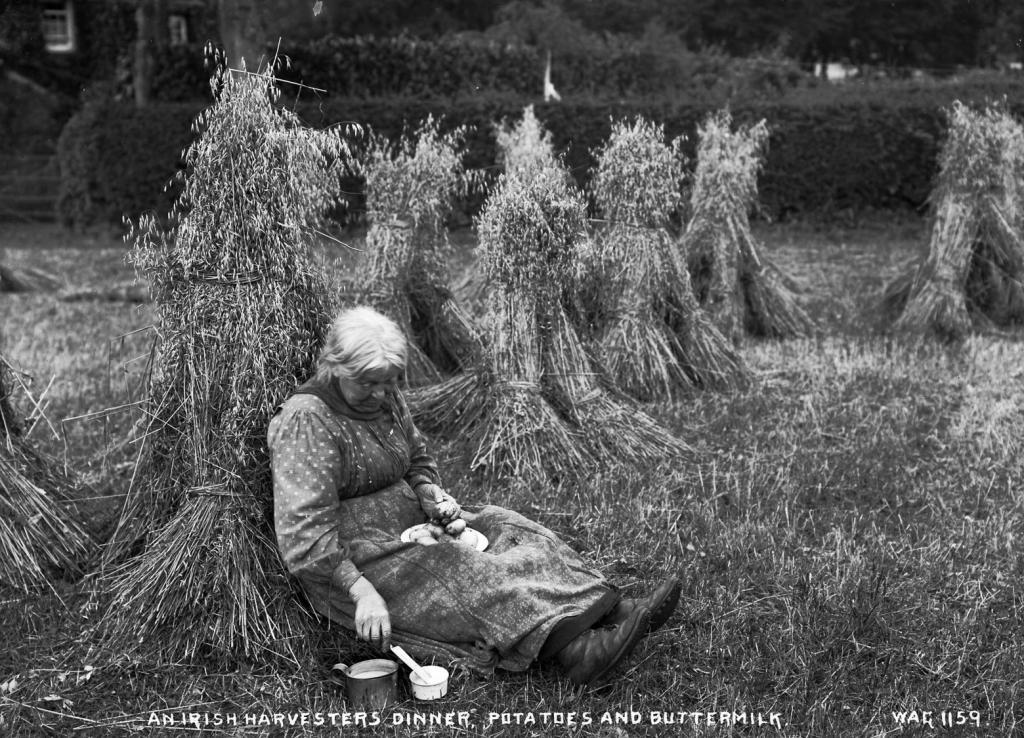 An old lady lying against a corn stack having a meal of potatoes and buttermilk