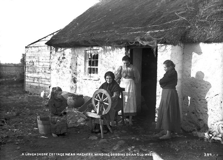 A woman outside a thatched cottage with spinning wheel and three girls