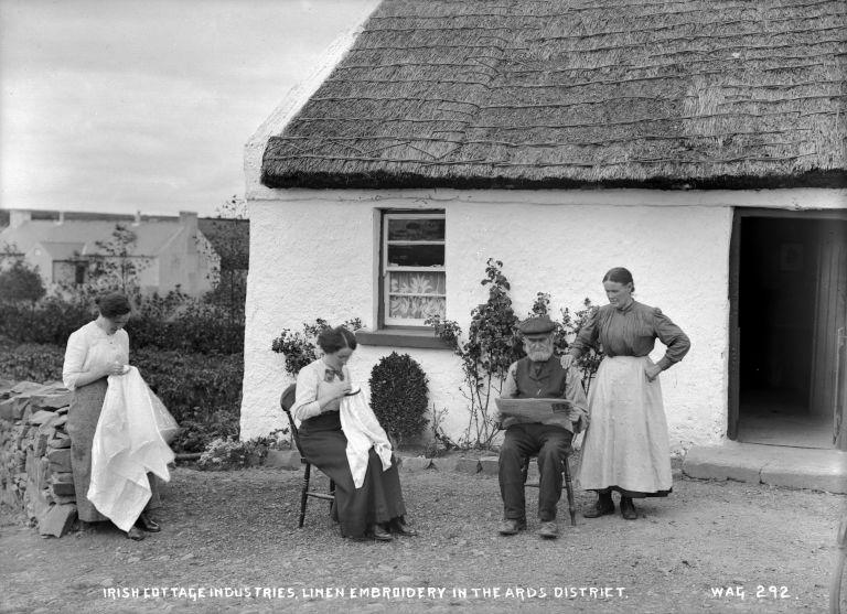 A group of people outside a thatched cottage in the Ards district, two are embroidering