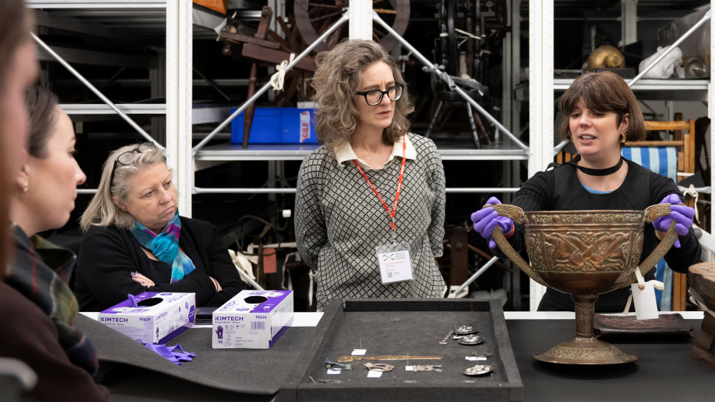 A group of people inside a museum store, the storage racks are in the background. The curator is talking to the group about an object that looks like a big celtic trophy, she's wearing purple gloves handling it. There's also a tray of silver artefacts in view on the table in front of them.