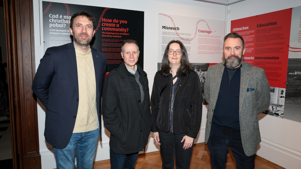 Four people standing in front of red, black and white exhibition panels mounted onto white walls. Two of them have their hands behind their backs, one has their hands in their pockets and the fourth person has their hands by their side. 