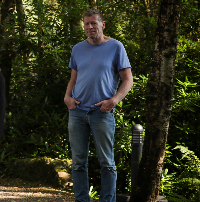 A curator from the Ulster American Folk Park in Omagh standing on a sunlit path outside in the Folk Park, with trees and shrubbery in the background. He is wearing a blue t-shirt and jeans and has his hands in his pockets. 