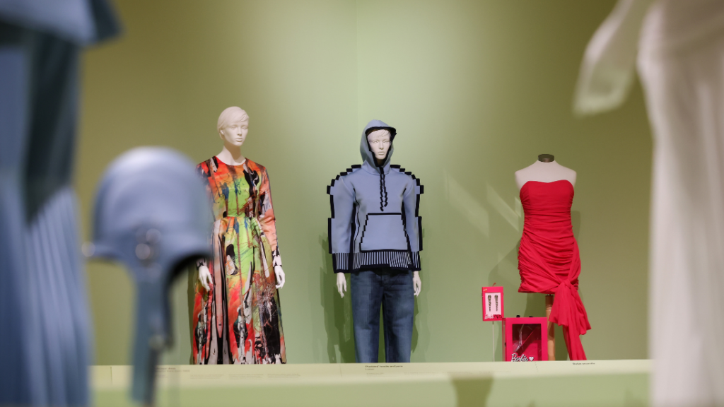 Three garments on white mannequins against a mint green wall. The object on the left is a colourful dress, the object in the middle is a blue, pixelated-looking hoodie and the object on the right is a pink dress