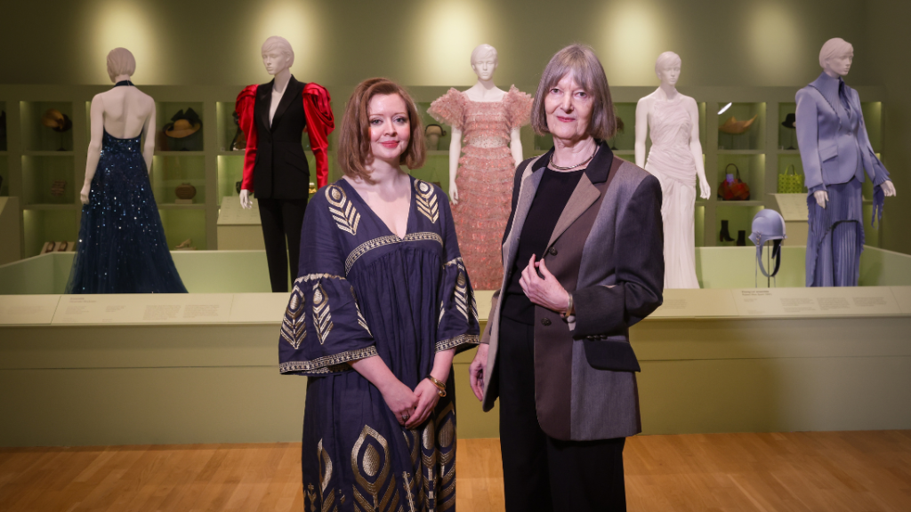 Two ladies standing in front of a display for fashion garments, with accessories in boxes on the back wall behind that. The lady on the left wears a dress and has shoulder length hair, the lady on the right is wearing a suit jacket and trousers and has collar bone length hair.