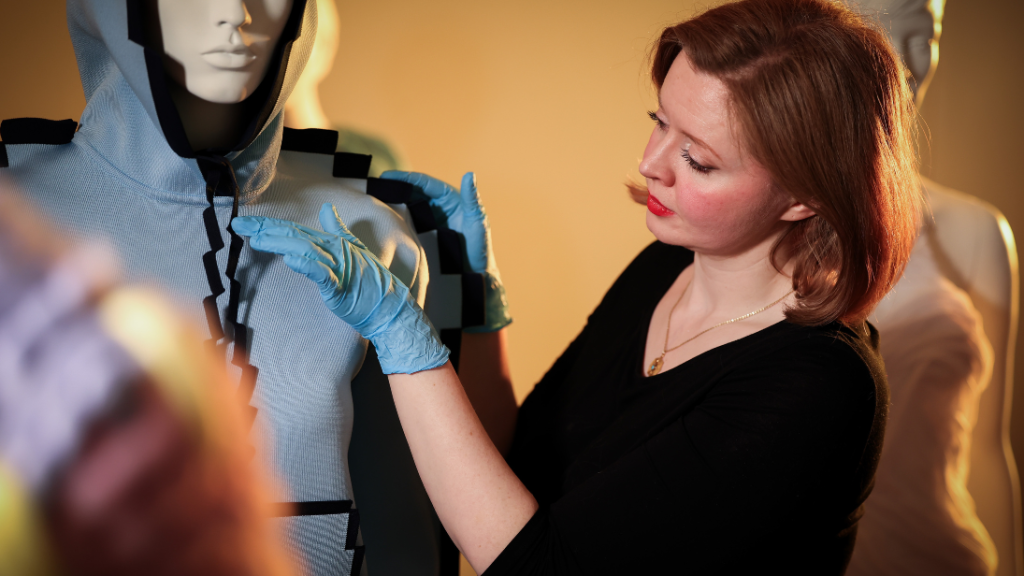 An art curator dressed in a black top wearing blue gloves carefully handles a museum object, which is a pixelated-effect blue and black hoodie by JW Anderson for Loewe. It is on a white mannequin and in shot behind the curator is another mannequin modelling an all white draped dress. The curator stands side on and is looking at the object in question, appearing to fix the hoodie.