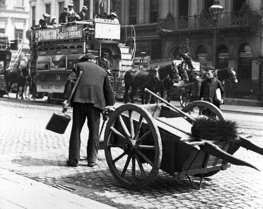 A man with a cart holding a big brush is in the foreground of this black and white photograph. In the background is a horse-drawn tram, which the man is cleaning up after.