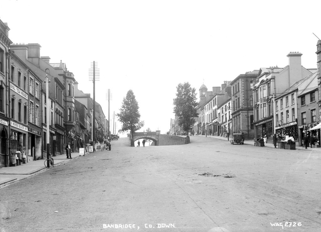 A black and white photograph of a town with a wide street. At the told of a small hill towards the end of the street, a small brick bridge sits across the road.