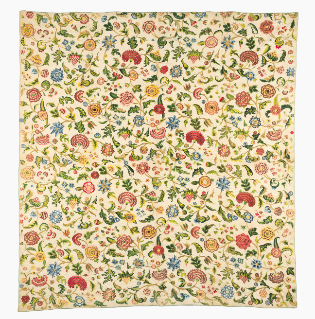 A large quilt. The background of the quilt is a charming yellow, with intricate floral embroidery across the entire quilt.