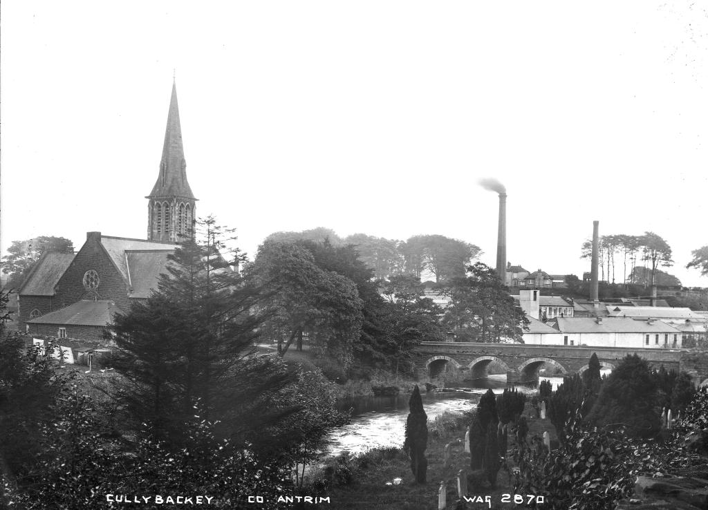 A black and white photograph of a town taken from afar, a church spire and a factory rising about some trees.