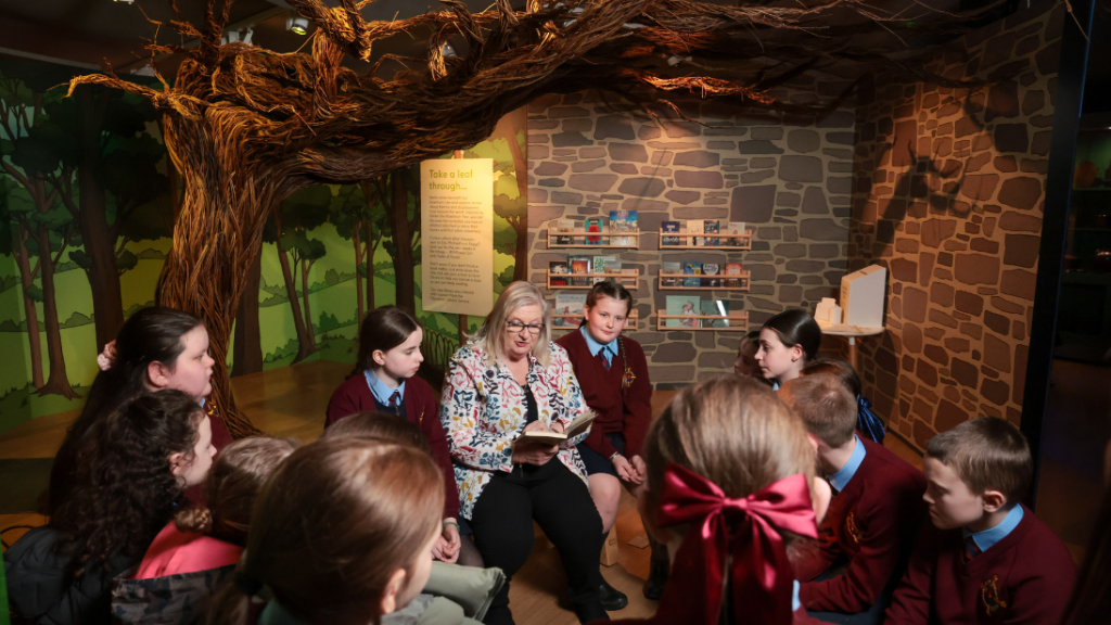 A group of school children in uniforms huddled around an author reading from her book, Under the Hawthorn Tree. They're inside an exhibition space and under a tree handmade from willow, which is the centrepiece to the exhibition.