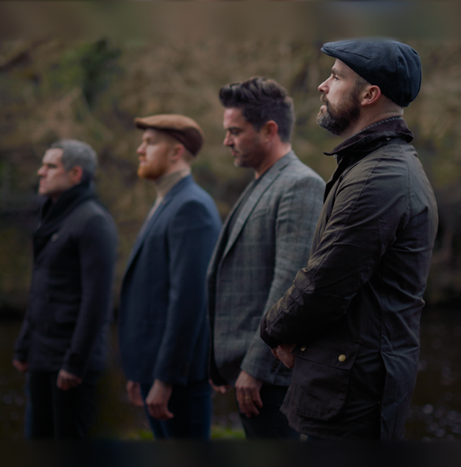 Four men standing in a line wearing autumn coats and two of the men are wearing flat caps