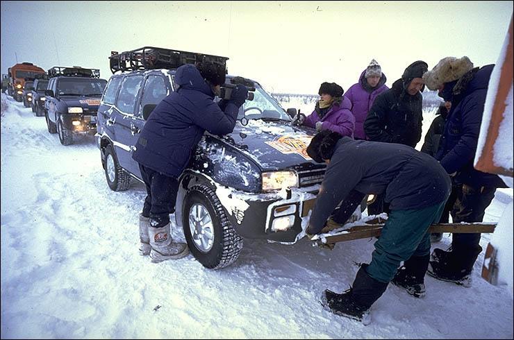 A photograph of a line of cars on a snowy road. At the front, a man is working of a car grille, while behind him, a young woman speak to a person holding a film camera.